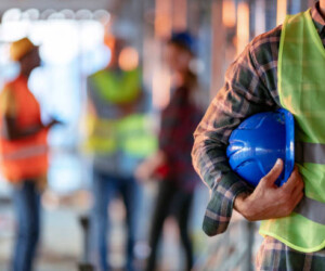 Man holding blue helmet close up. Construction man worker with office and people in background. Close up of a construction worker's hand holding working helmet.